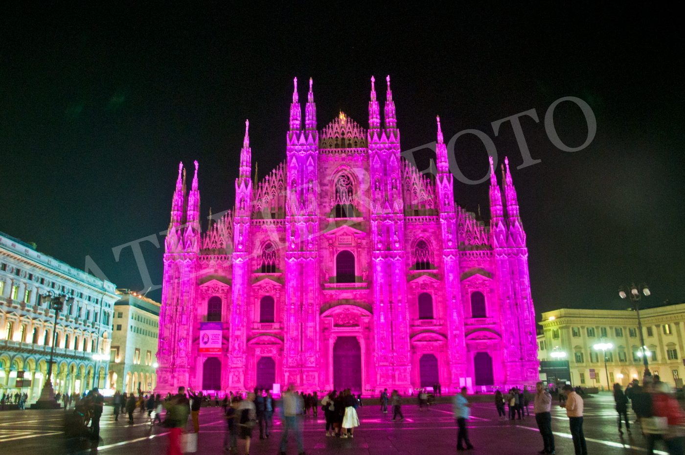 Veduta notturna frontale del Duomo di Milano illuminato di rosa, con persone in movimento nella piazza.