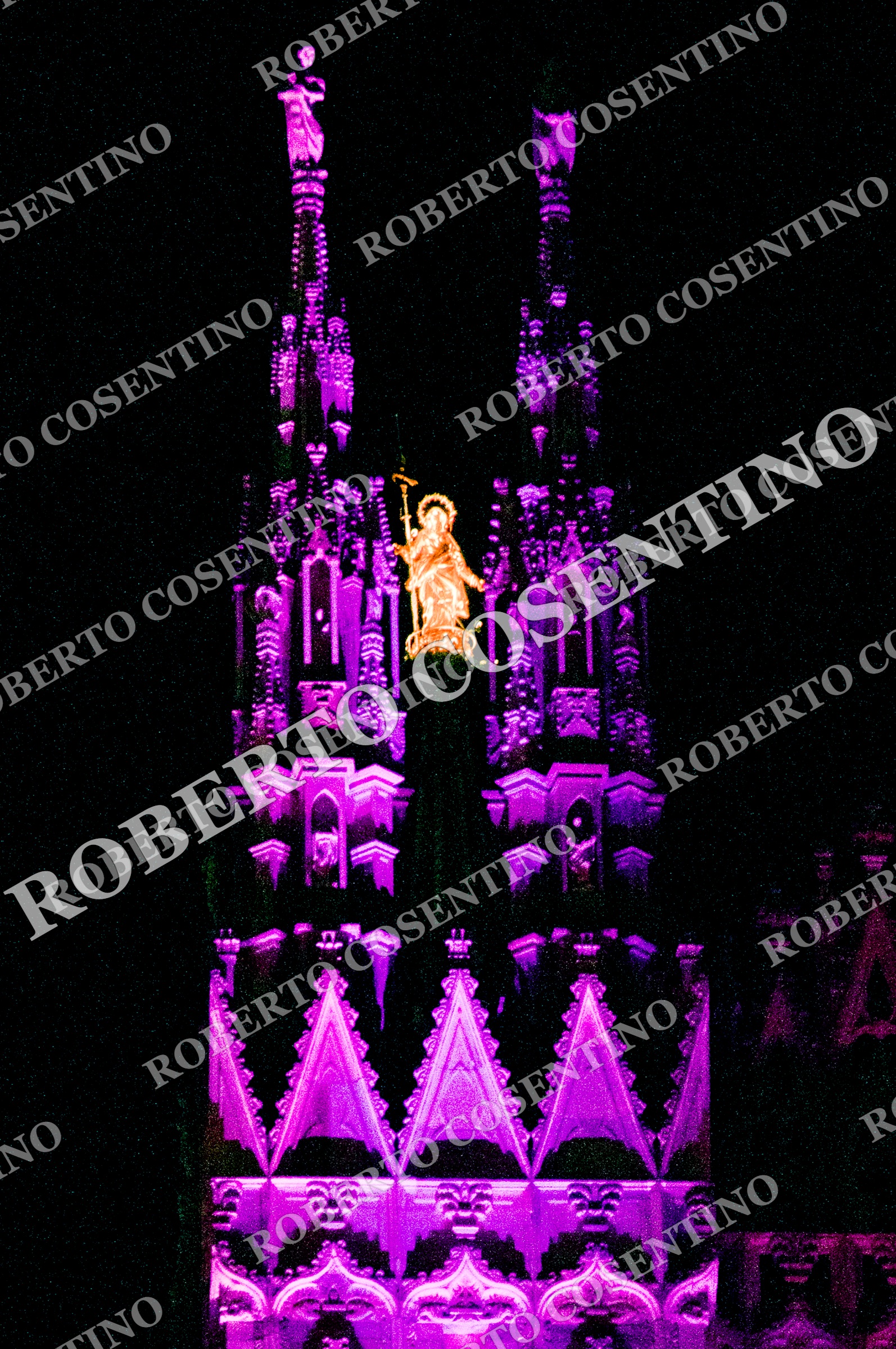 Dettaglio notturno delle guglie del Duomo di Milano illuminate di viola, con la Madonnina dorata al centro.