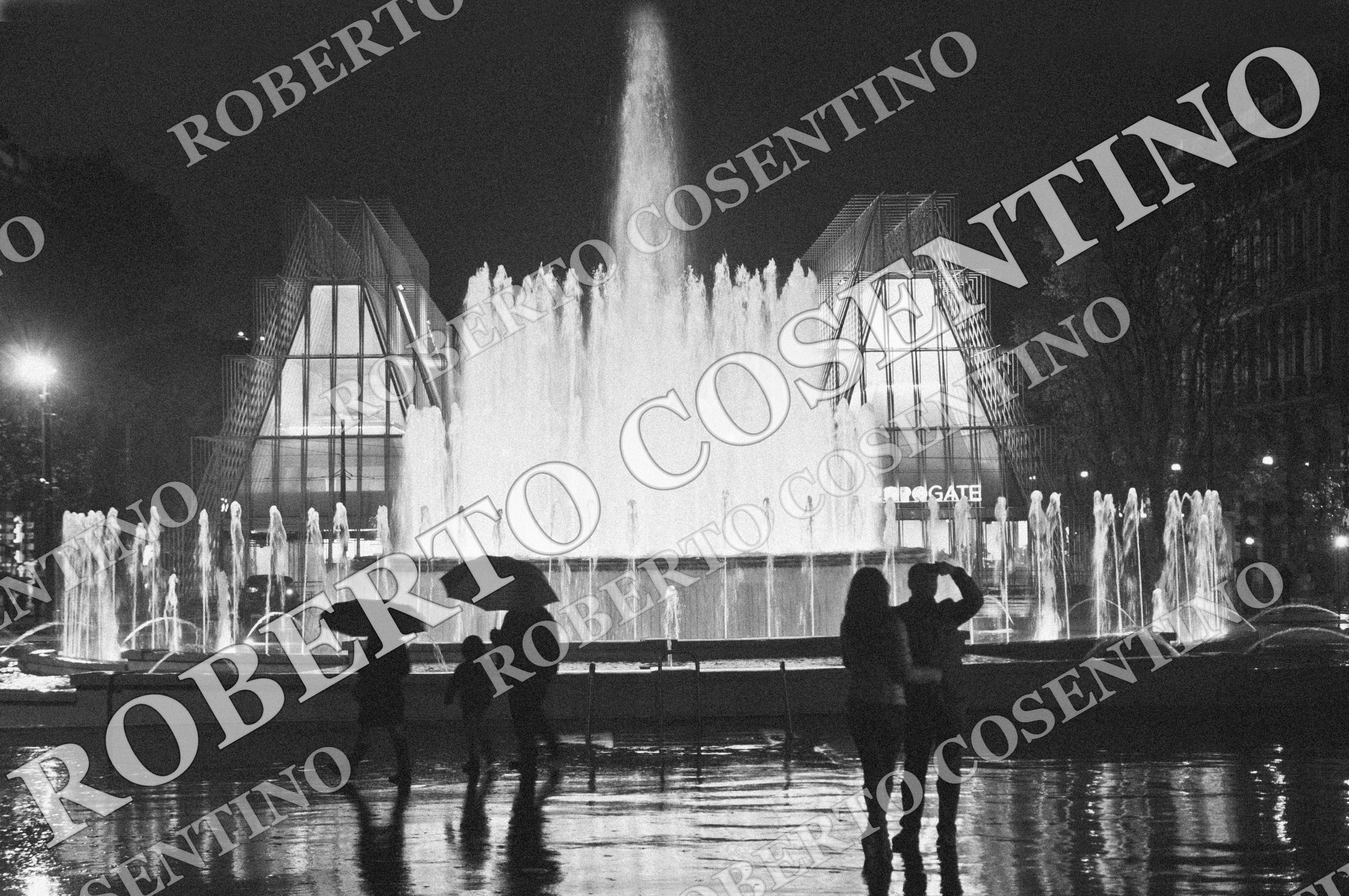 Fotografia in bianco e nero della fontana illuminata di Piazza Castello a Milano, con persone sotto gli ombrelli e riflessi sul pavimento bagnato.